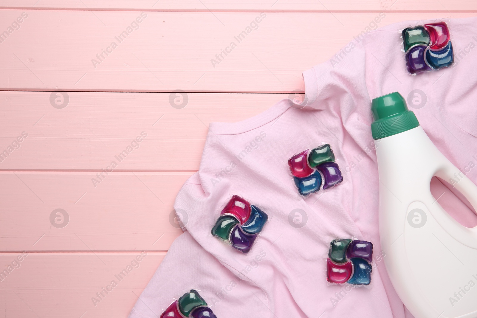 Photo of Laundry detergent in bottle, pods and t-shirt on pink wooden table, top view. Space for text