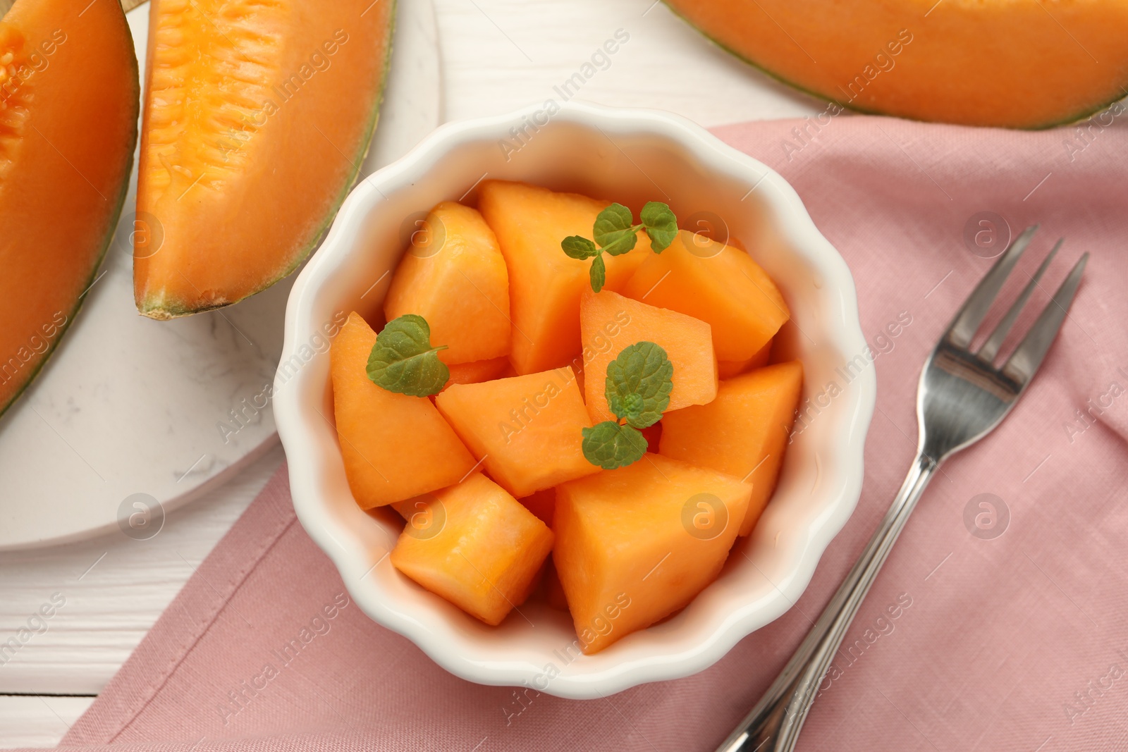 Photo of Pieces of ripe Cantaloupe melon in bowl and fork on white wooden table, flat lay