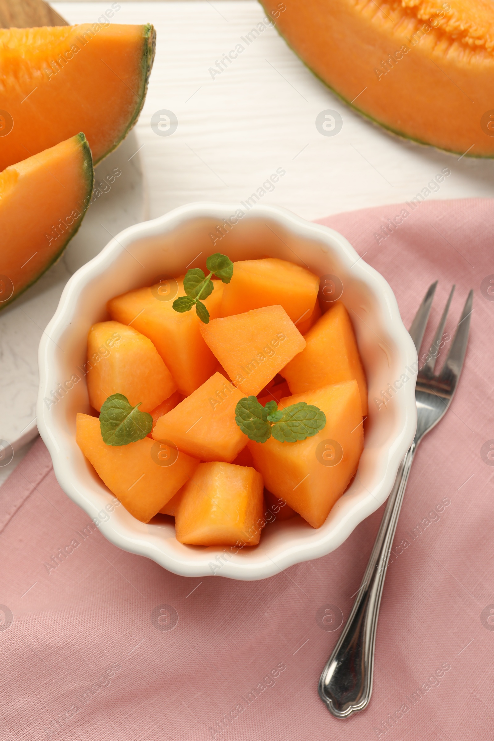 Photo of Pieces of ripe Cantaloupe melon in bowl and fork on white wooden table, flat lay