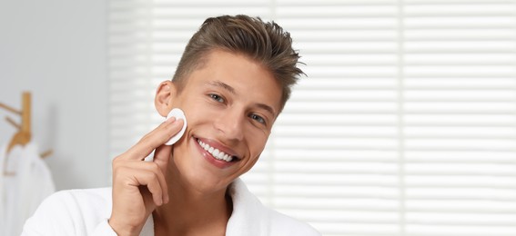 Handsome young man cleaning face with cotton pad in bathroom. Banner design with space for text