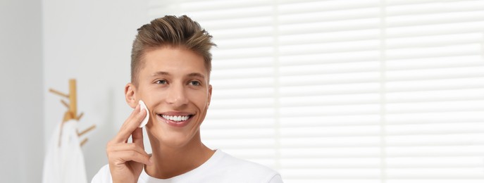 Image of Handsome young man cleaning face with cotton pad in bathroom. Banner design with space for text