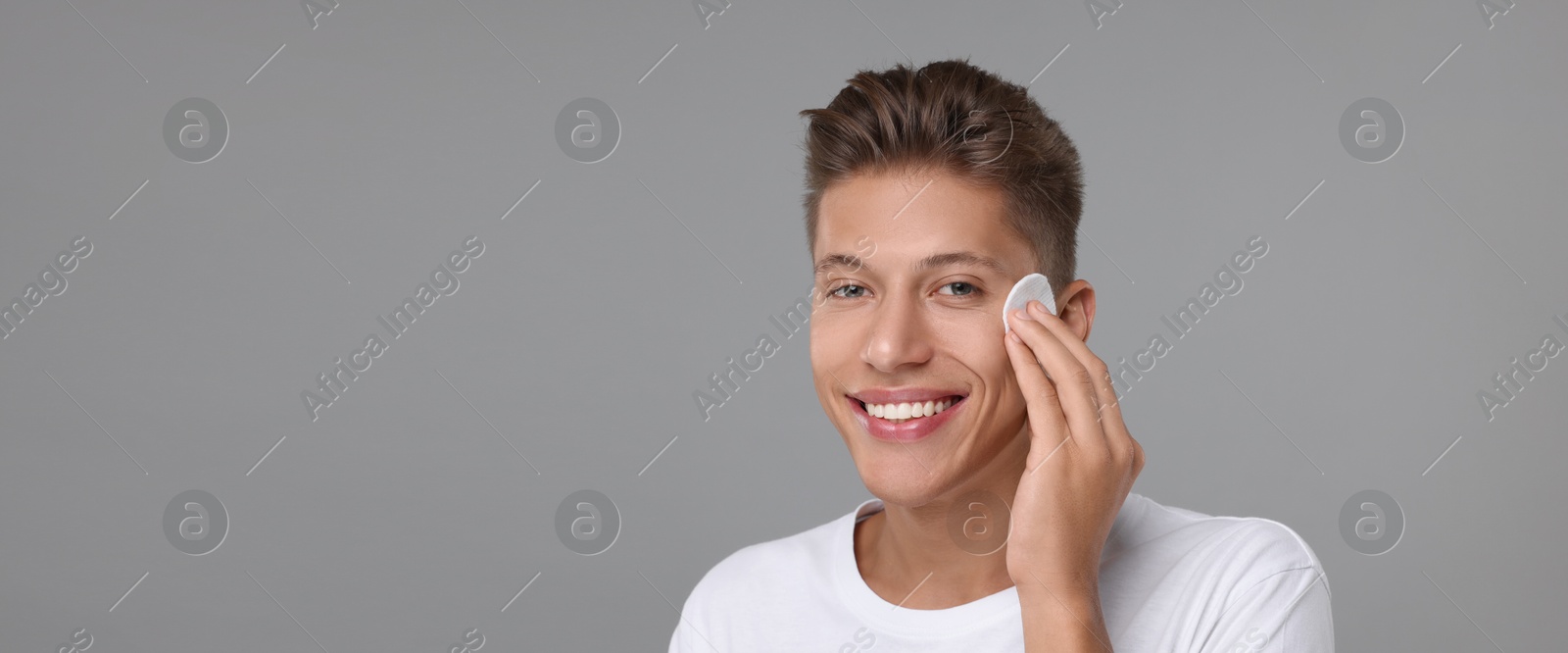 Image of Handsome young man cleaning face with cotton pad on grey background. Banner design with space for text