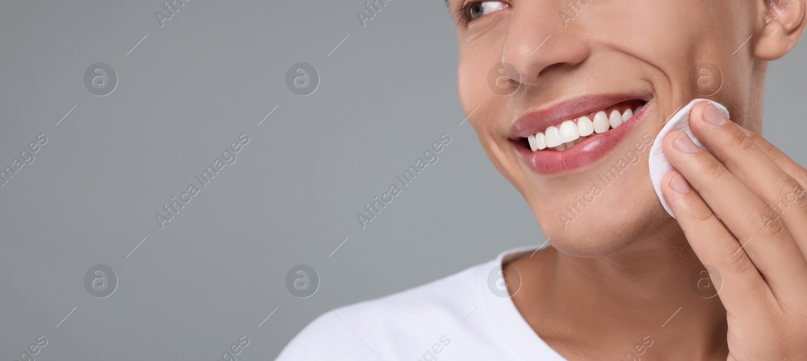 Image of Young man cleaning face with cotton pad on grey background, closeup. Banner design with space for text