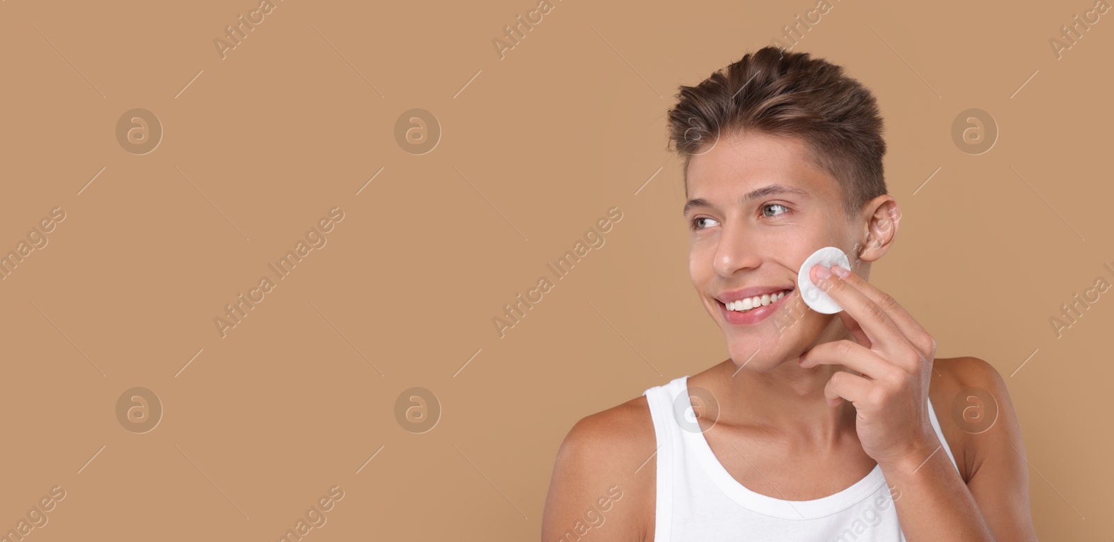 Image of Handsome young man cleaning face with cotton pad on dark beige background. Banner design with space for text