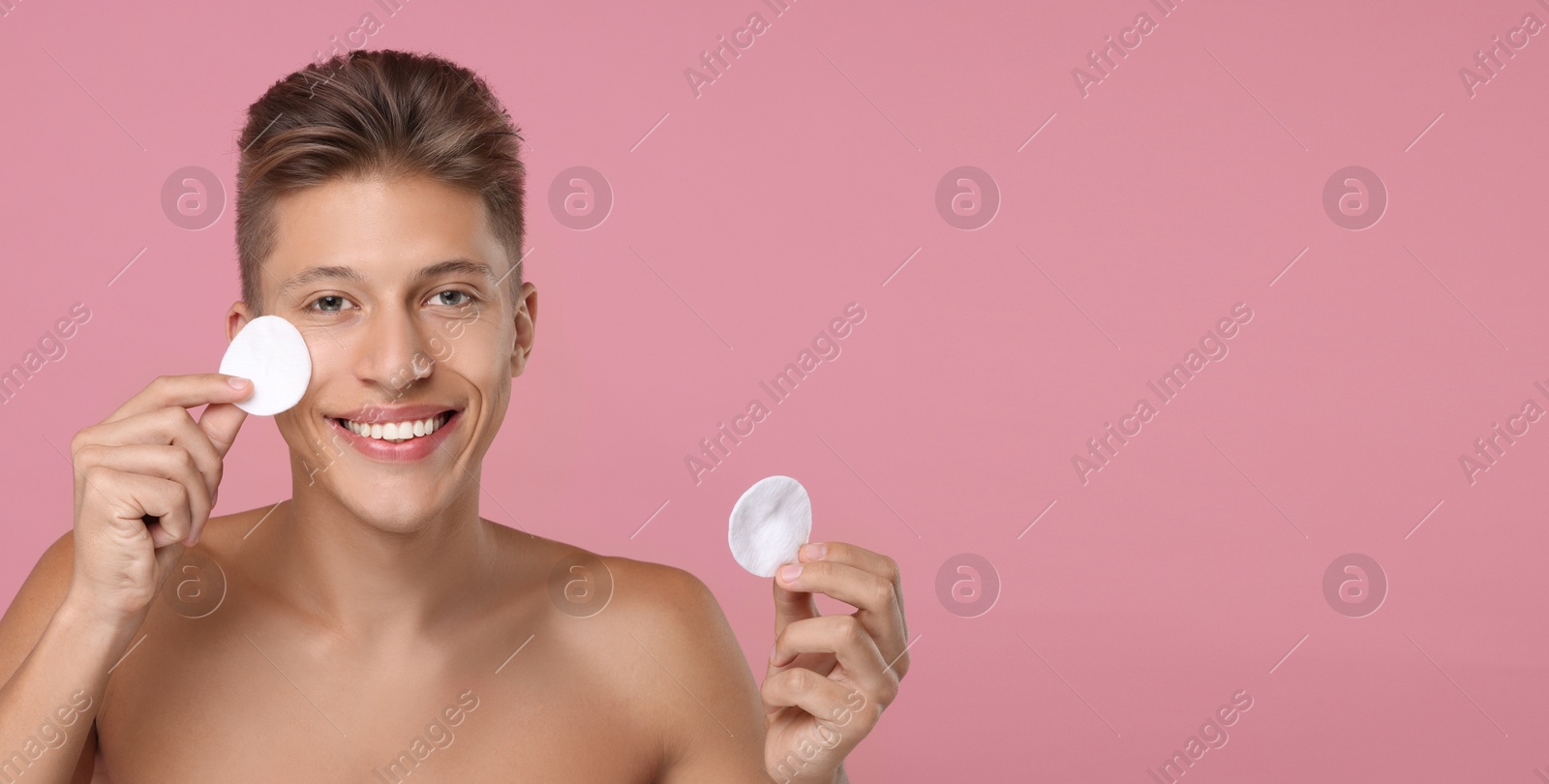 Image of Handsome young man cleaning face with cotton pads on pink background. Banner design with space for text