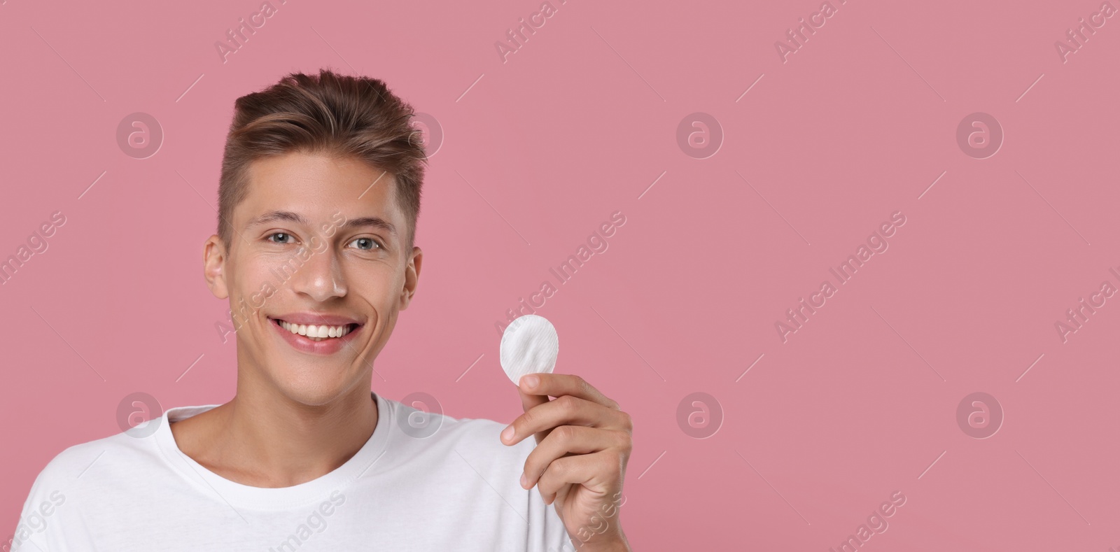 Image of Handsome young man cleaning face with cotton pad on pink background. Banner design with space for text