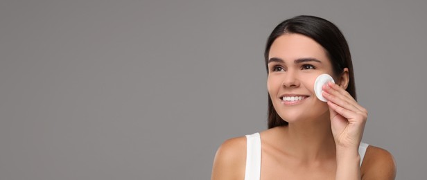 Young woman cleaning her face with cotton pad on grey background. Banner design with space for text