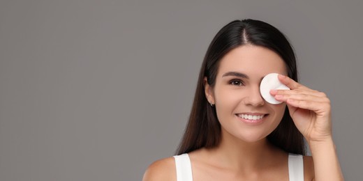 Young woman cleaning her face with cotton pad on grey background. Banner design with space for text