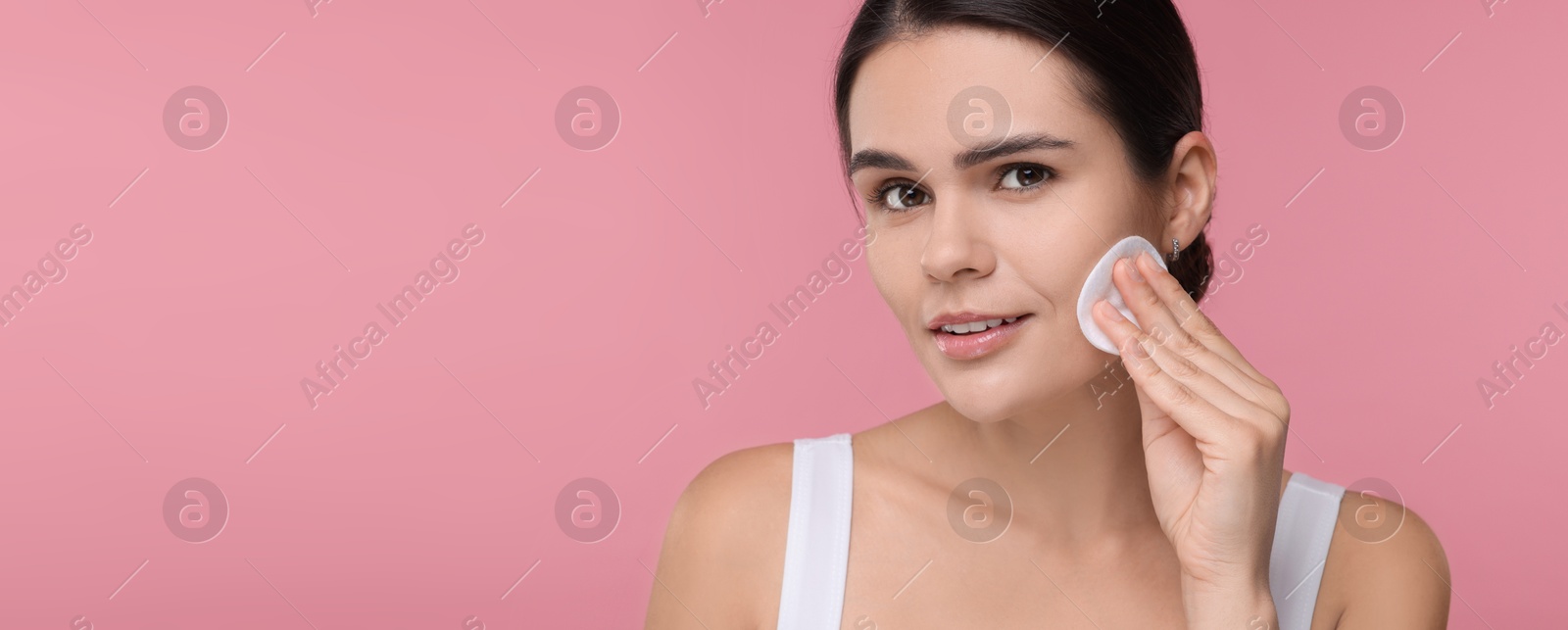 Image of Young woman cleaning her face with cotton pad on pink background. Banner design with space for text