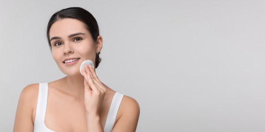 Image of Young woman cleaning her face with cotton pad on light grey background. Banner design with space for text