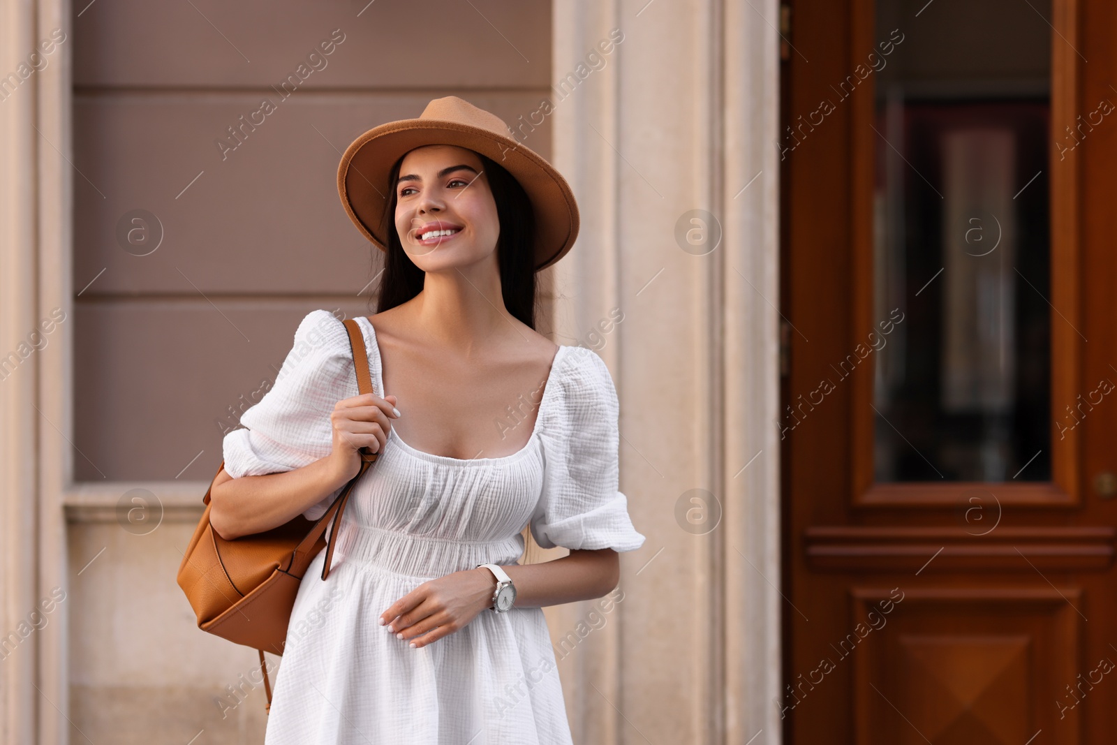 Photo of Beautiful young woman in stylish hat with backpack on city street, space for text