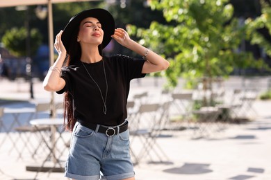 Photo of Beautiful young woman in stylish black hat on city street, space for text