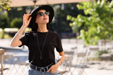 Photo of Young woman in stylish black hat and sunglasses on city street, space for text