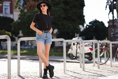 Photo of Young woman in stylish black hat and sunglasses on city street, space for text