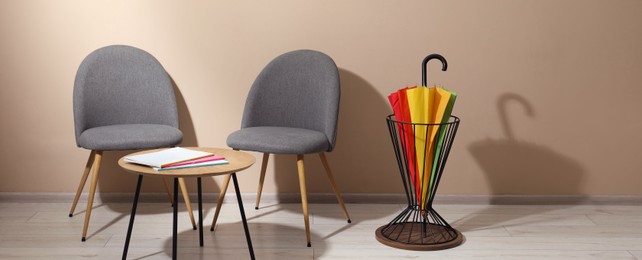 Bright umbrella in holder, coffee table and chairs near beige wall. Space for text