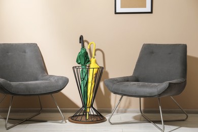 Photo of Bright umbrellas in holder and armchairs near beige wall