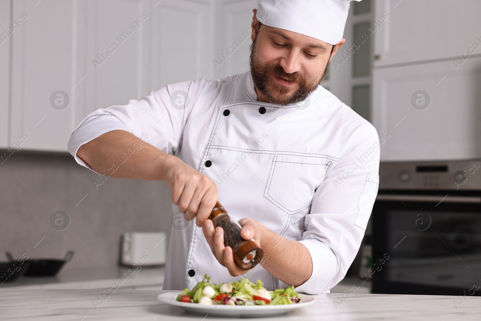 Photo of Professional chef adding pepper to delicious salad at white marble table in kitchen