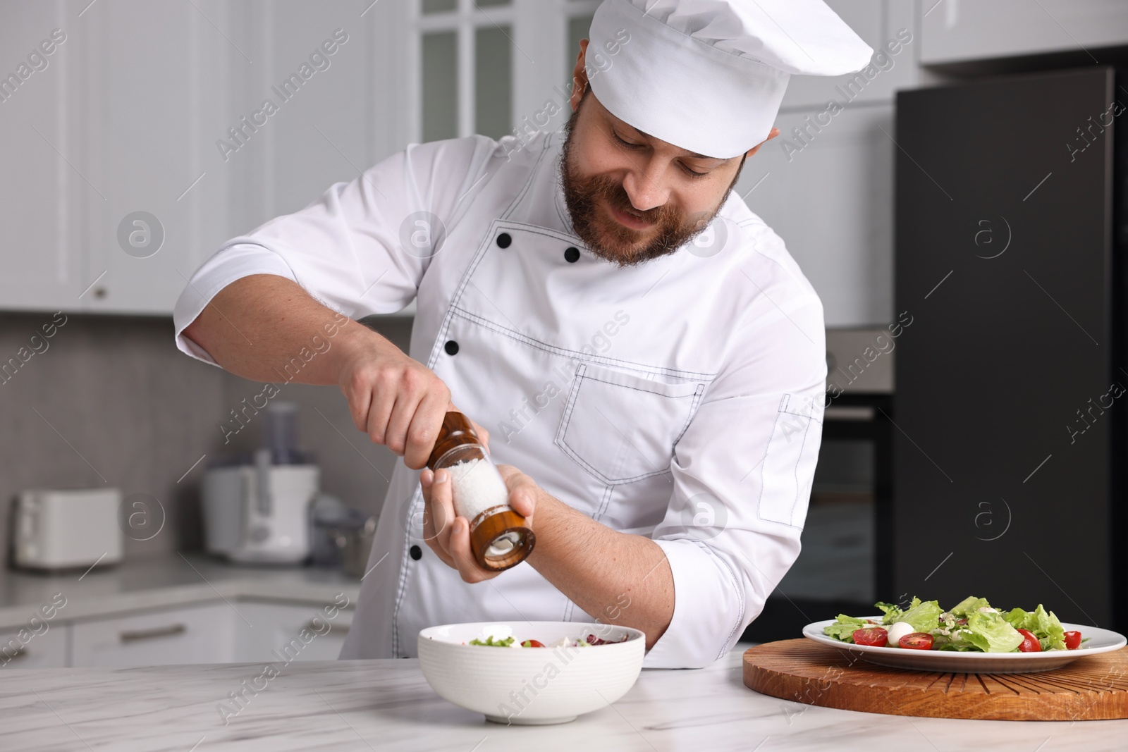 Photo of Professional chef adding salt into bowl with salad at white marble table in kitchen