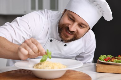 Photo of Professional chef decorating delicious pasta with basil at white marble table in kitchen