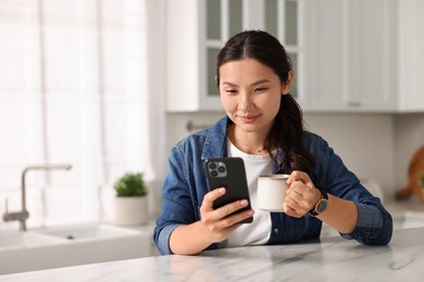 Photo of Beautiful woman with smartphone drinking coffee at table in kitchen. Space for text