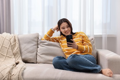 Photo of Smiling woman using smartphone on sofa at home