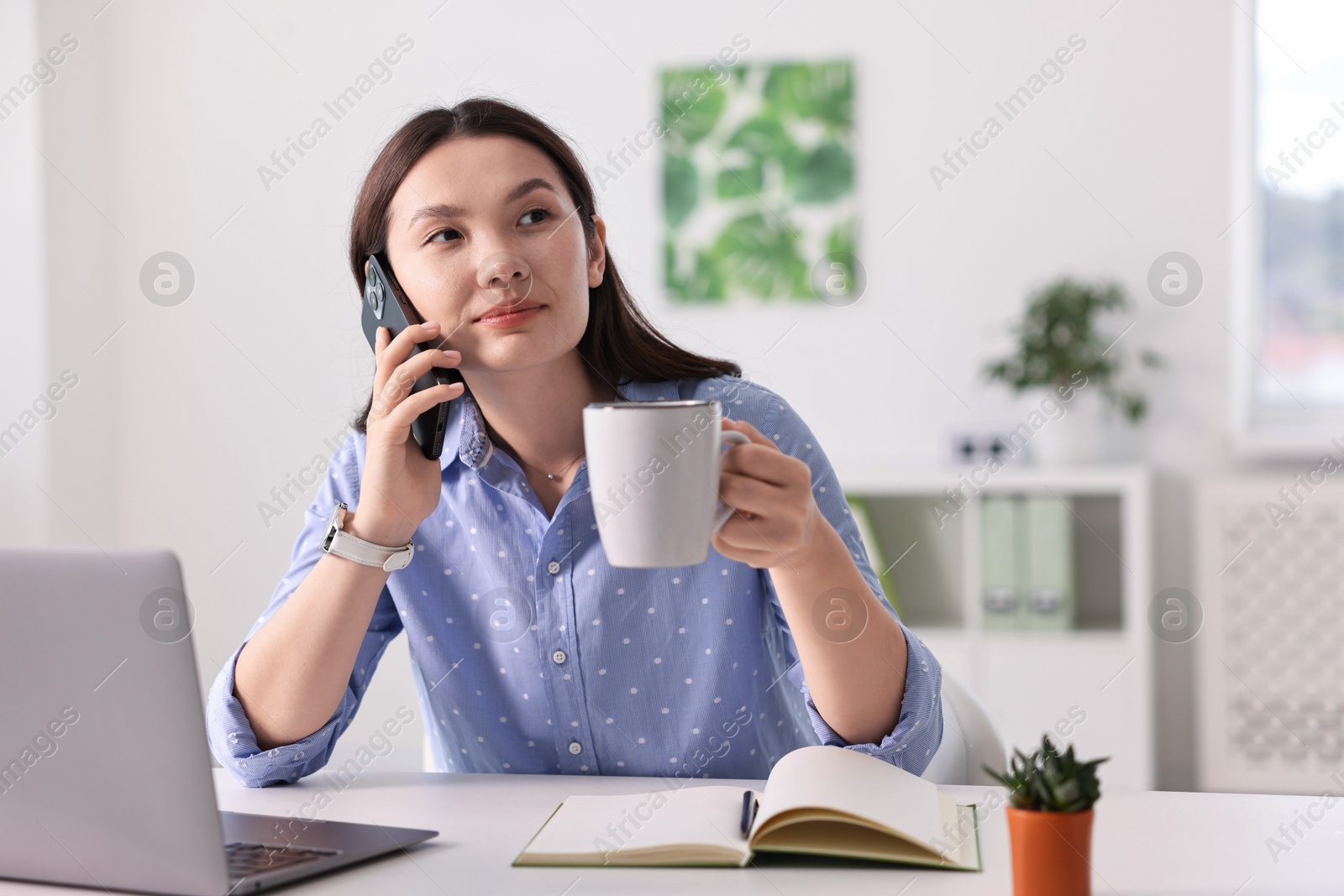 Photo of Beautiful businesswoman with cup talking by smartphone at table in office