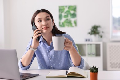 Photo of Beautiful businesswoman with cup talking by smartphone at table in office