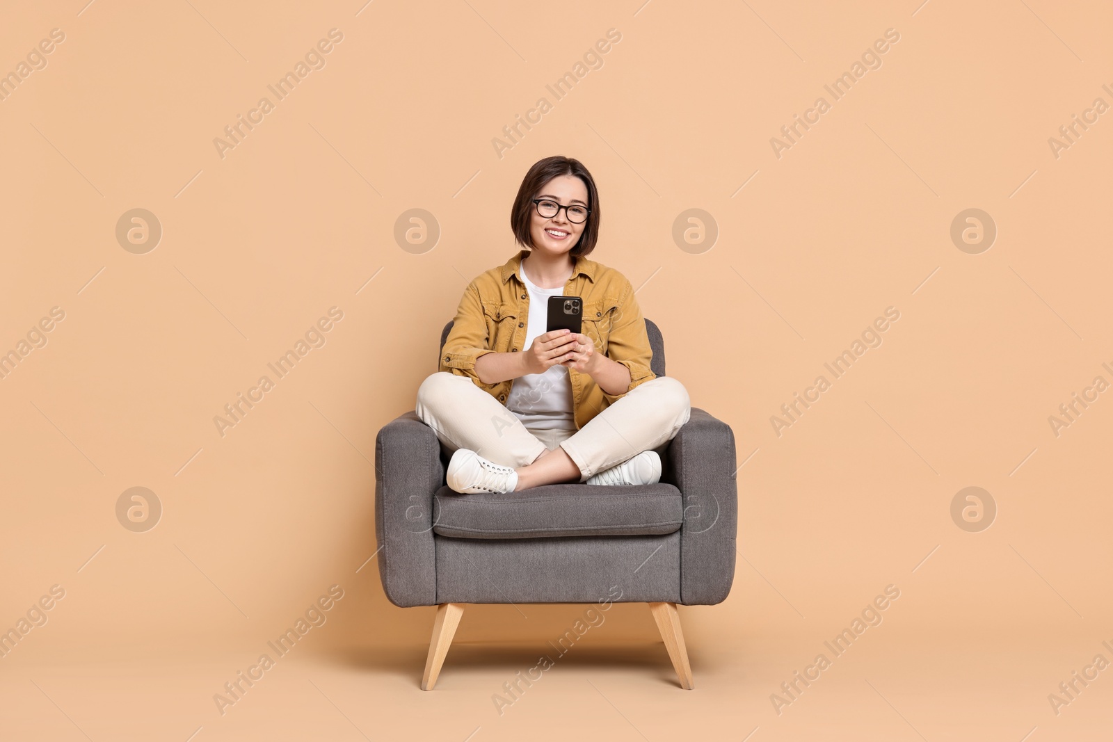 Photo of Smiling woman with smartphone sitting on armchair against beige background