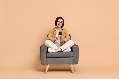 Smiling woman with smartphone sitting on armchair against beige background