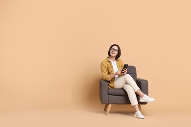 Smiling woman with smartphone sitting on armchair against beige background. Space for text