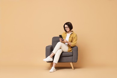 Photo of Smiling woman with smartphone sitting on armchair against beige background