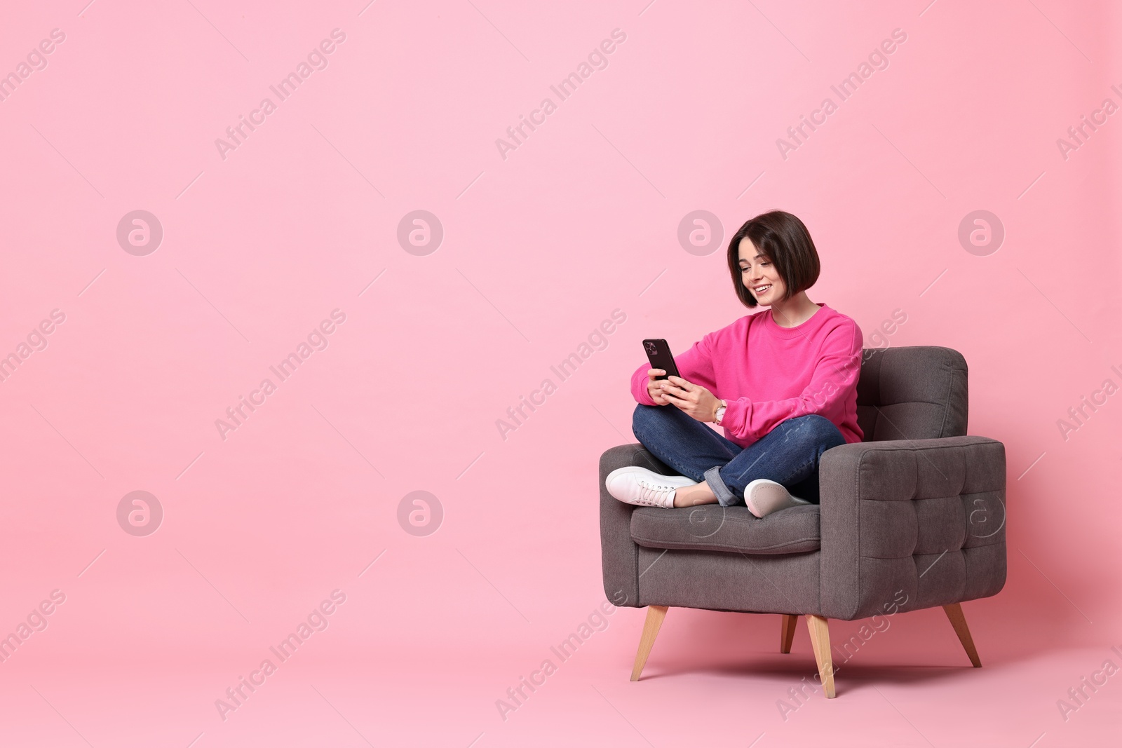 Photo of Smiling woman with smartphone sitting on armchair against pink background. Space for text