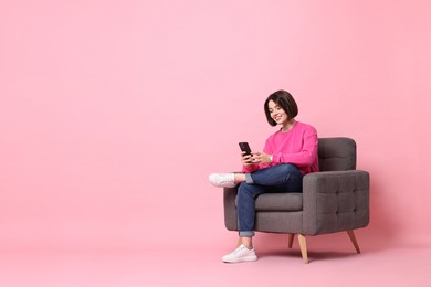 Photo of Smiling woman with smartphone sitting on armchair against pink background. Space for text