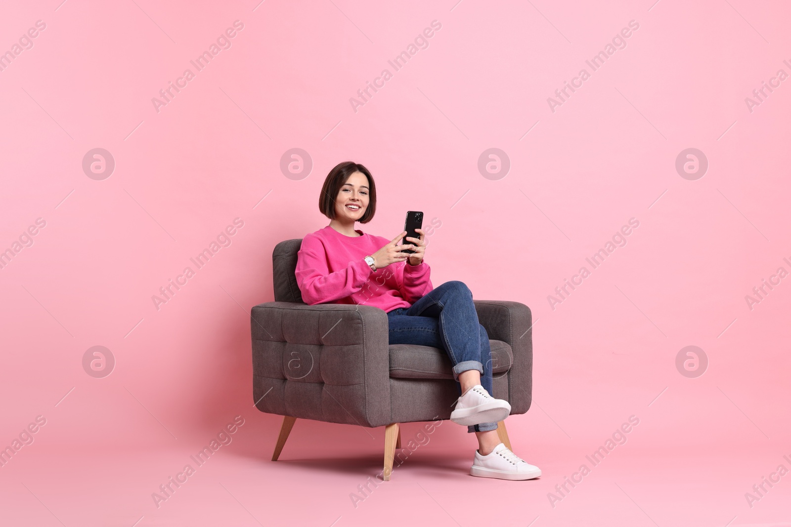 Photo of Smiling woman with smartphone sitting on armchair against pink background