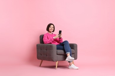 Smiling woman with smartphone sitting on armchair against pink background