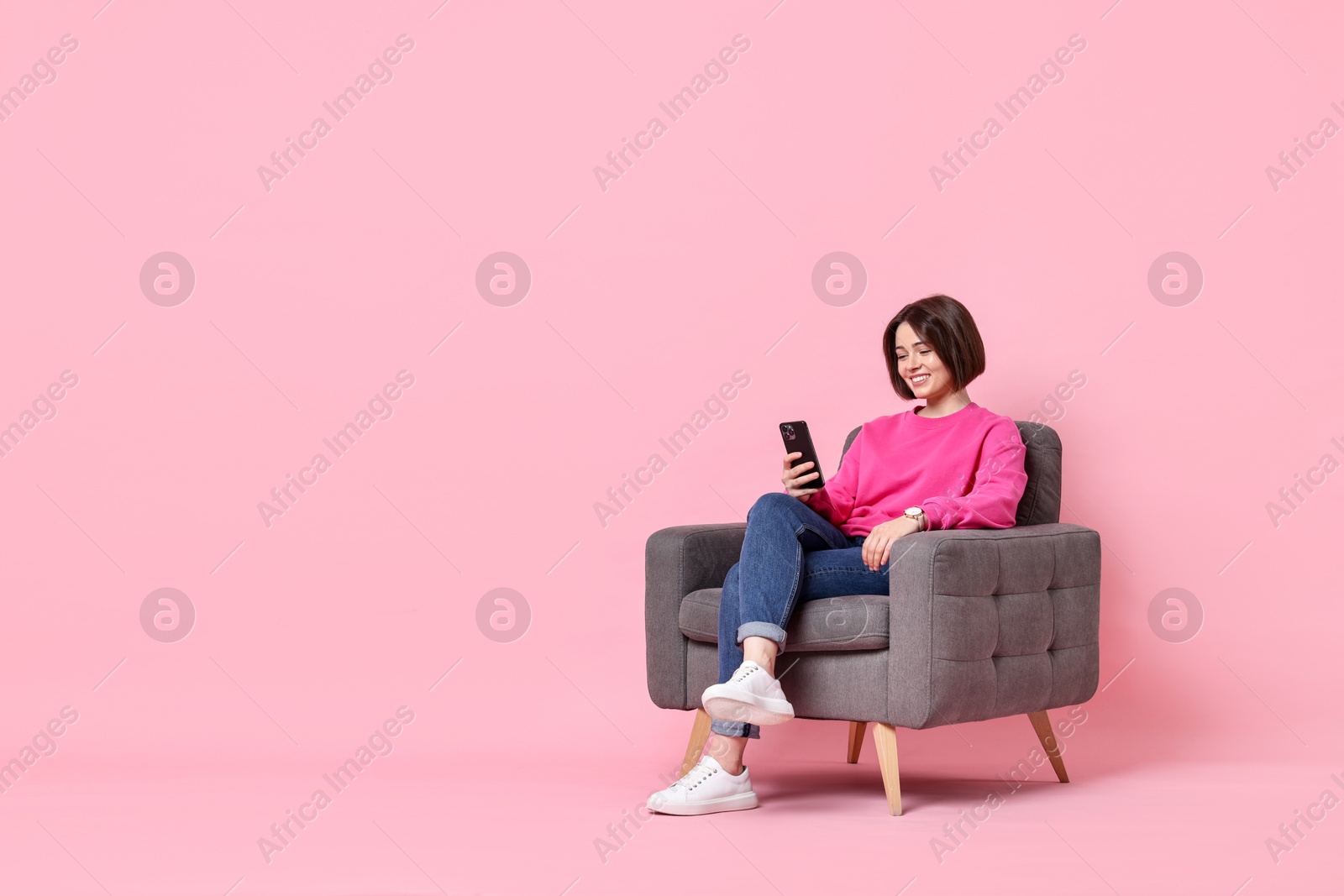 Photo of Smiling woman with smartphone sitting on armchair against pink background. Space for text