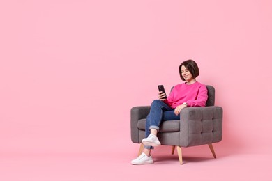 Smiling woman with smartphone sitting on armchair against pink background. Space for text