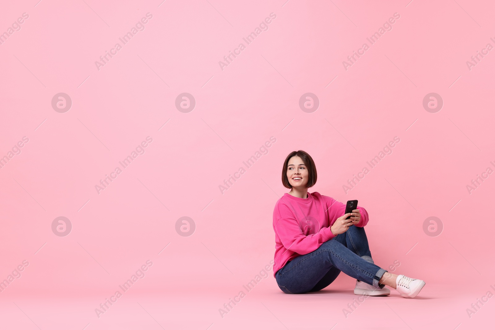 Photo of Smiling woman with smartphone sitting on pink background. Space for text