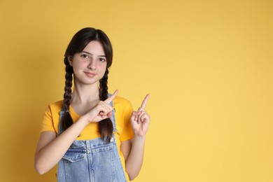 Portrait of cute teenage girl pointing at something on yellow background. Space for text