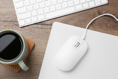 Photo of Computer mouse with mousepad, cup of coffee and keyboard on wooden table, flat lay