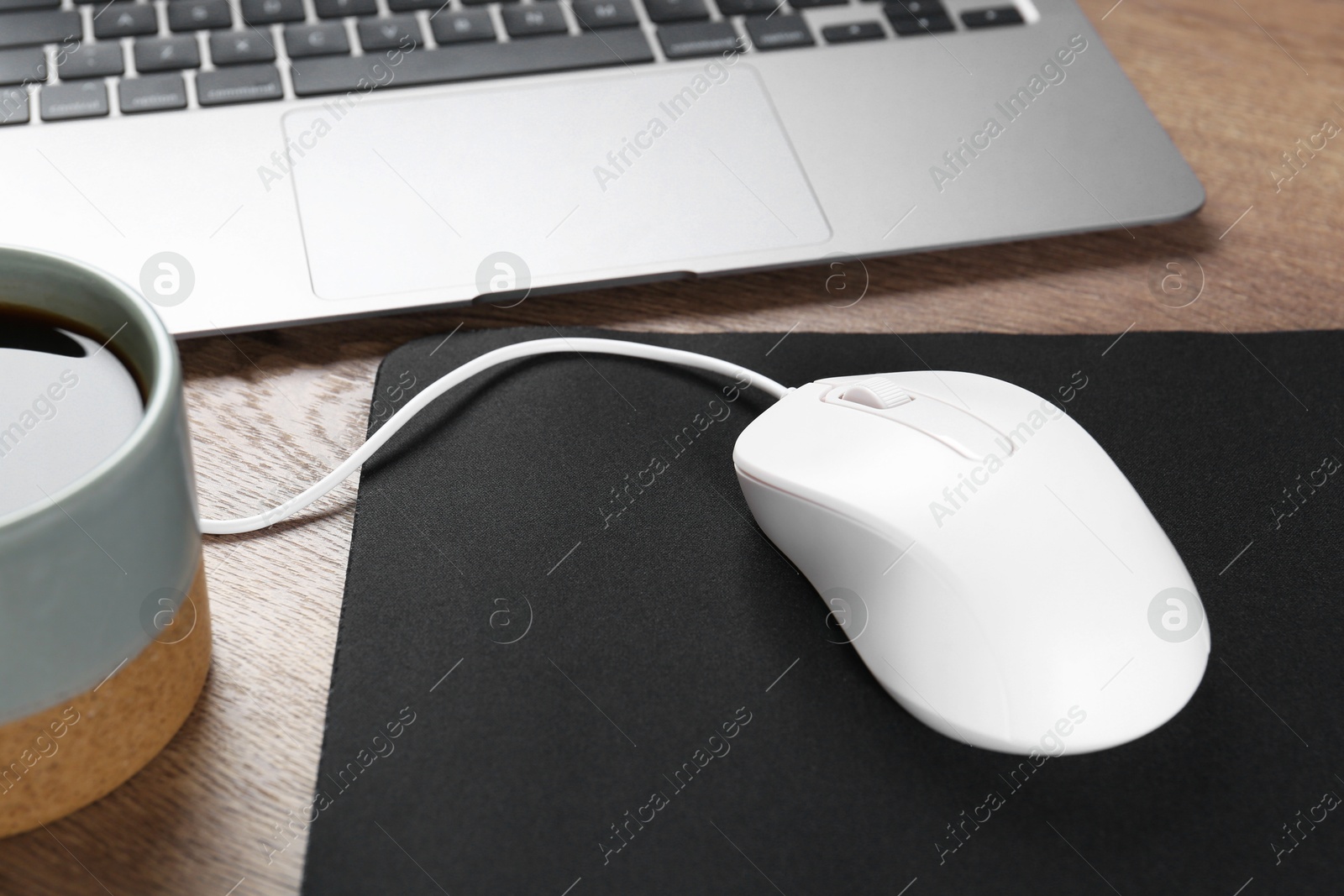 Photo of Computer mouse with mousepad, cup of coffee and laptop on wooden table