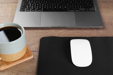 Photo of Computer mouse with mousepad, cup of coffee and laptop on wooden table