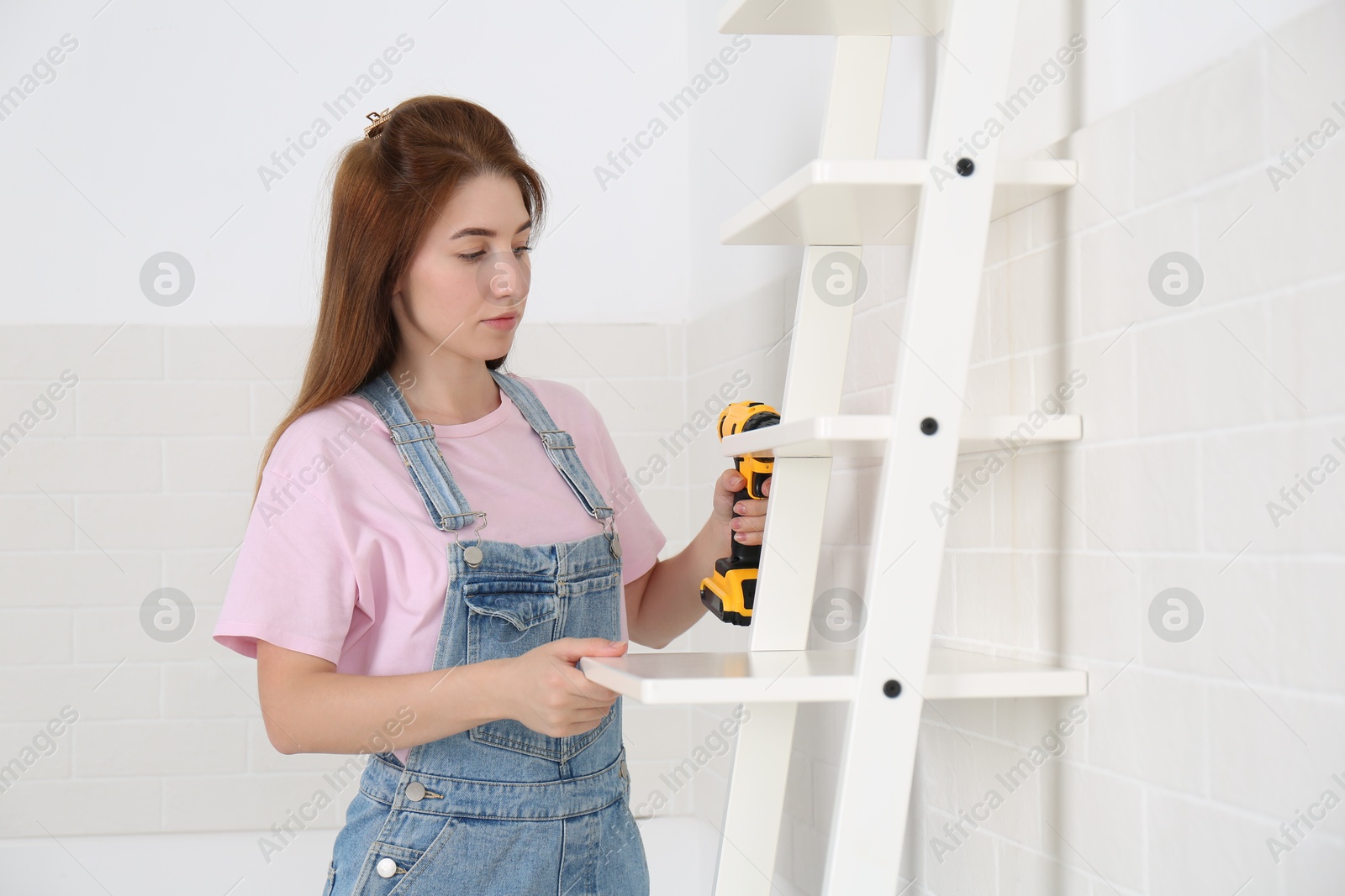 Photo of Woman with electric screwdriver assembling ladder shelf indoors