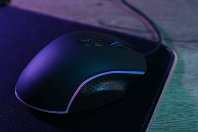 Photo of Black computer mouse and mousepad on wooden table in neon lights, closeup