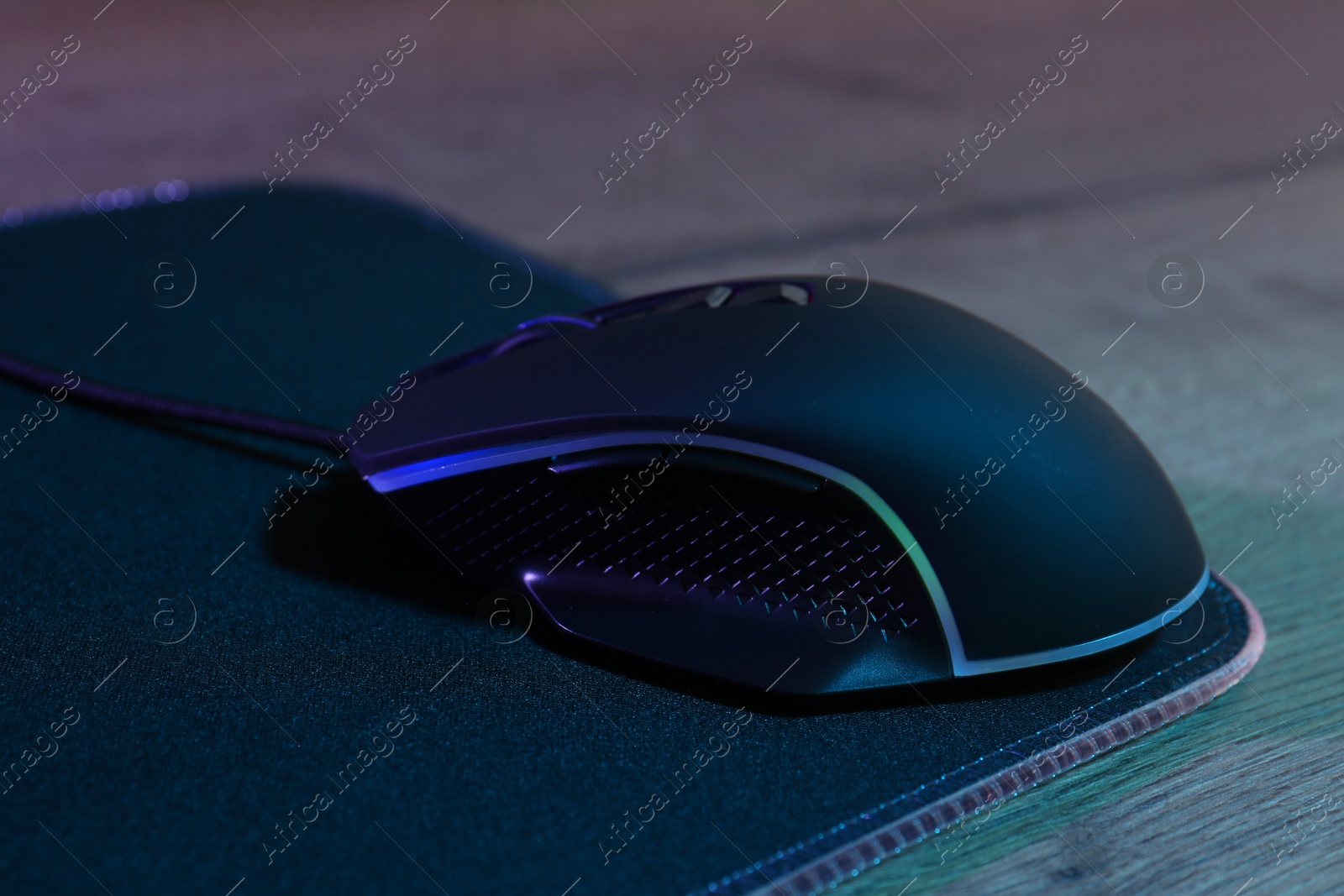 Photo of Black computer mouse and mousepad on wooden table in neon lights, closeup