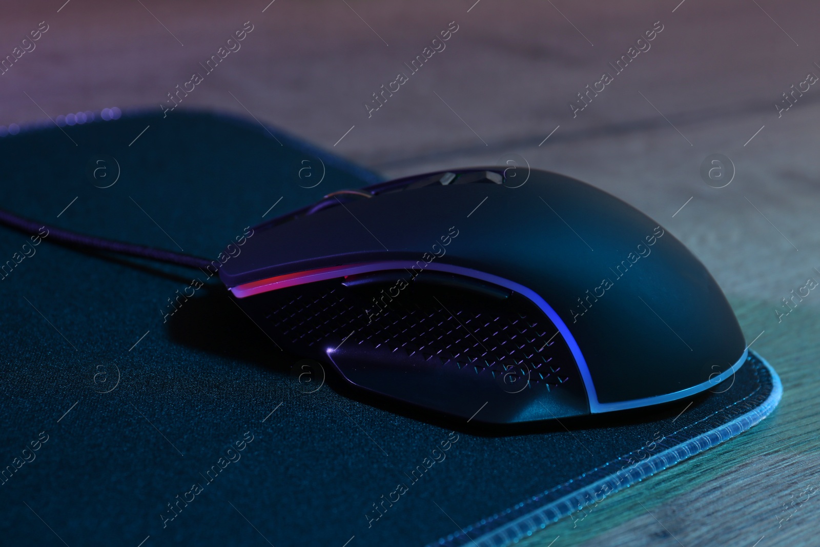 Photo of Black computer mouse and mousepad on wooden table in neon lights, closeup