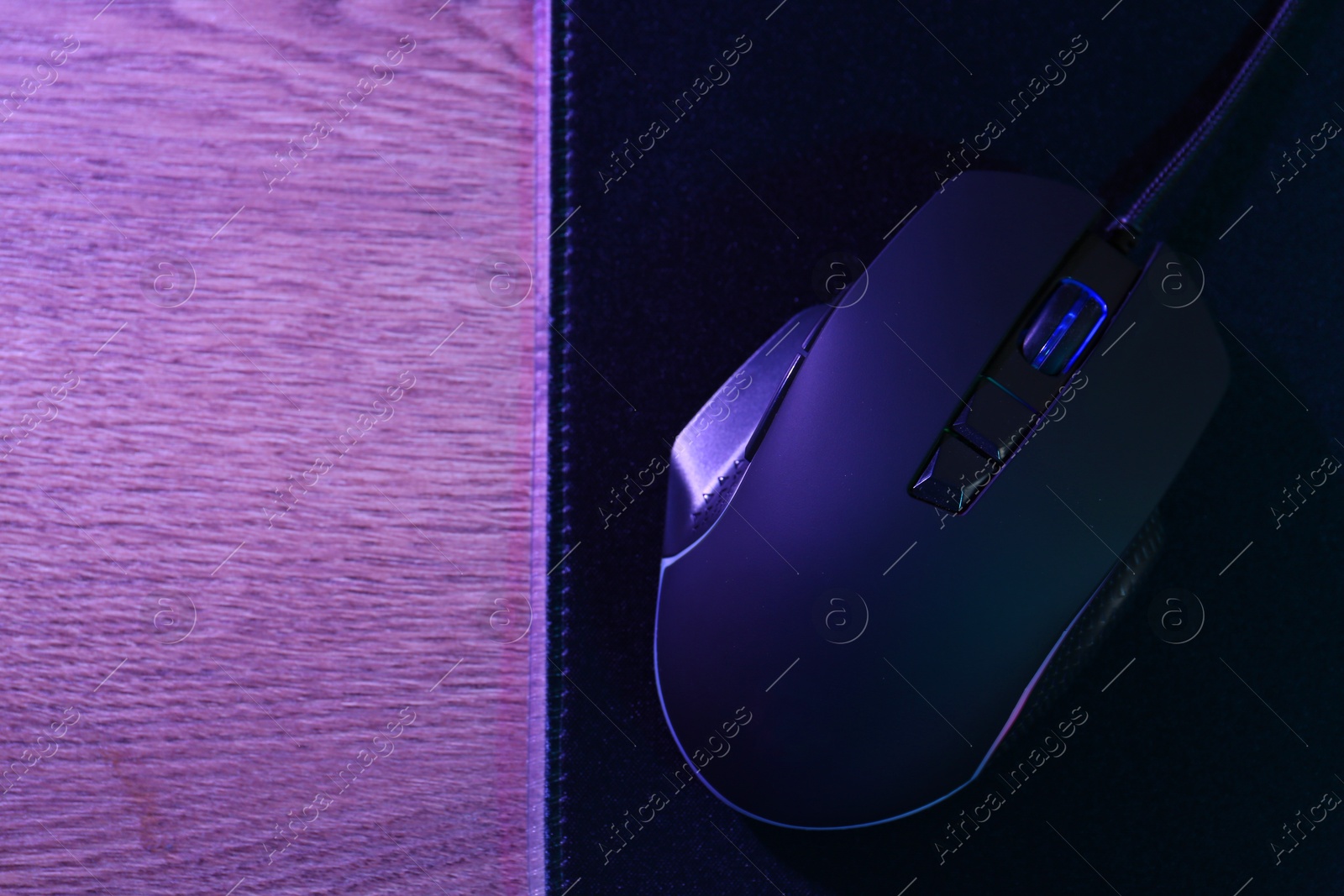 Photo of Black computer mouse and mousepad on wooden table in neon lights, top view. Space for text