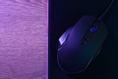 Photo of Black computer mouse and mousepad on wooden table in neon lights, top view. Space for text