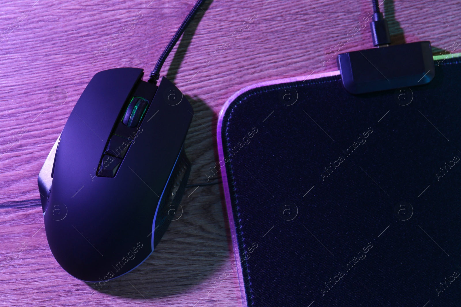 Photo of Black computer mouse and mousepad on wooden table in neon lights, top view