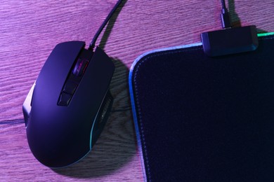 Photo of Black computer mouse and mousepad on wooden table in neon lights, top view